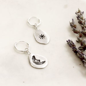 May include: A pair of silver hoop earrings featuring dangling charms. One charm is shaped like a teardrop with a cut-out sun design, while the other charm is a teardrop shape with a cut-out crescent moon design, both with a polished finish.