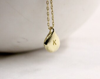 May include: A gold-colored necklace featuring a delicate chain and a small, smooth, teardrop-shaped pendant. The pendant has a polished, reflective surface and is stamped with the uppercase letter K in a simple, clean font on its front face.