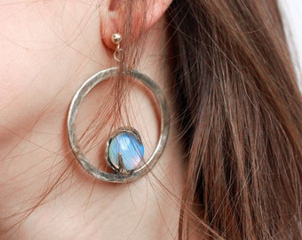 May include: A silver-toned hoop earring featuring a textured, hammered metal finish. A single, iridescent, round cabochon stone with blue and white hues is set within the lower portion of the circular frame, secured by a small metal prong setting.
