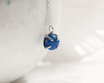May include: A silver chain necklace featuring a round, deep blue lapis lazuli gemstone pendant. The stone is secured in a four-prong claw setting that grips the rounded surface of the gem. The delicate metal chain connects to the top of the setting.