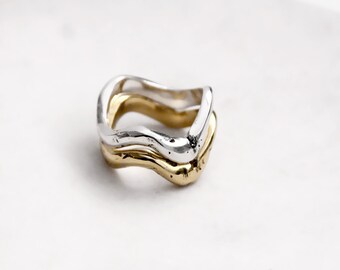 May include: A two-tone ring featuring a stacked design with one silver-colored band and one gold-colored band. Both bands have an irregular, wavy, and organic shape with a smooth, polished finish, creating a layered appearance on the finger.