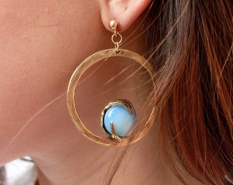 May include: A gold-toned hoop earring featuring a textured circular frame with a small, smooth, light blue spherical stone held in place by a claw-like setting at the bottom. The earring attaches to the earlobe with a simple gold-toned stud post.