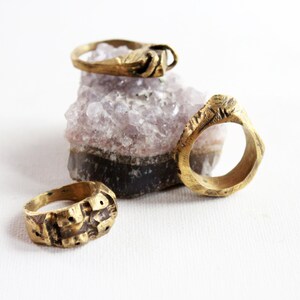 May include: Three gold-toned metal rings with textured, uneven surfaces and organic shapes. Two rings are positioned in the foreground, while one ring rests on top of a jagged, light purple and dark gray crystalline mineral cluster.