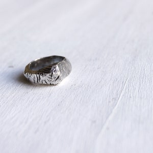 May include: A silver-toned ring featuring a textured, rugged surface with deep, irregular grooves and a polished metallic finish. The band has a thick, uneven profile with a distinct, organic appearance that catches the light across its ridges.