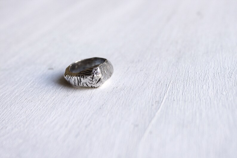 May include: A silver-toned ring featuring a textured, rugged surface with deep, irregular grooves and a polished metallic finish. The band has a thick, uneven profile with a distinct, organic appearance that catches the light across its ridges.