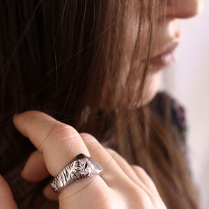 May include: A silver-toned ring featuring a textured, rugged surface with deep grooves and a raised, irregular top section. The band has a metallic finish and a distinct, uneven pattern that wraps around the finger, showcasing a raw, cast-metal appearance.