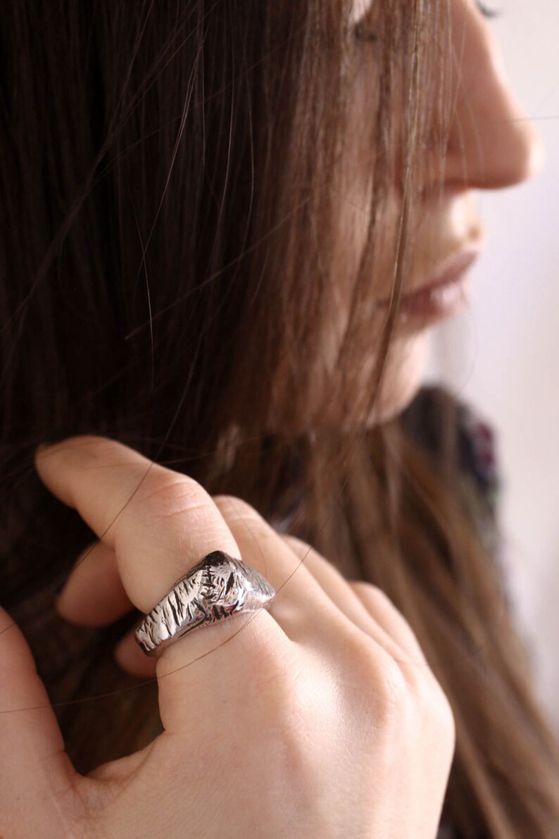 May include: A silver-toned ring featuring a textured, rugged surface with deep grooves and a raised, irregular top section. The band has a metallic finish and a distinct, uneven pattern that wraps around the finger, showcasing a raw, cast-metal appearance.