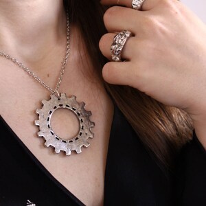 May include: A silver-toned necklace featuring a large gear-shaped pendant on a thin chain, accompanied by two silver-toned rings with a textured, uneven surface. The jewelry pieces have a metallic finish and a distinct industrial-inspired design.