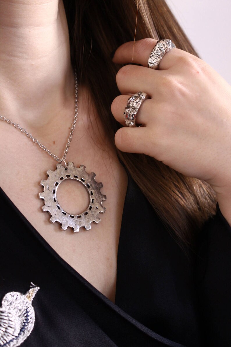 May include: A silver-toned necklace featuring a large gear-shaped pendant on a thin chain, accompanied by two silver-toned rings with a textured, uneven surface. The jewelry pieces have a metallic finish and a distinct industrial-inspired design.