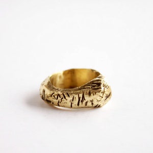 May include: A gold-colored ring with a thick, uneven band featuring a textured, bark-like surface. The metal has a matte finish with deep, irregular grooves carved into the exterior, creating a rugged, organic appearance around the entire circumference.