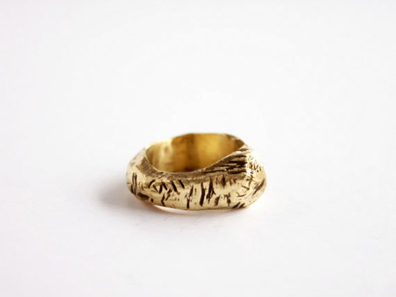 May include: A gold-colored ring with a thick, uneven band featuring a textured, bark-like surface. The metal has a matte finish with deep, irregular grooves carved into the exterior, creating a rugged, organic appearance around the entire circumference.