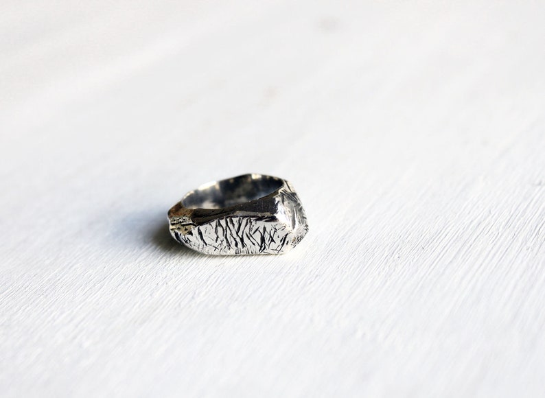 May include: A silver ring featuring a highly textured, irregular surface with deep, etched grooves and a metallic finish. The band has a thick, uneven profile that creates a rugged appearance, showcasing a distinct, hand-crafted, organic shape.