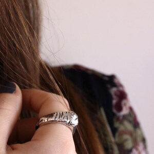 May include: A silver-toned ring featuring a textured, irregular surface with deep grooves and a polished finish. The band has a substantial, rounded profile that wraps around the finger, showcasing a rugged, organic aesthetic with visible metallic highlights.