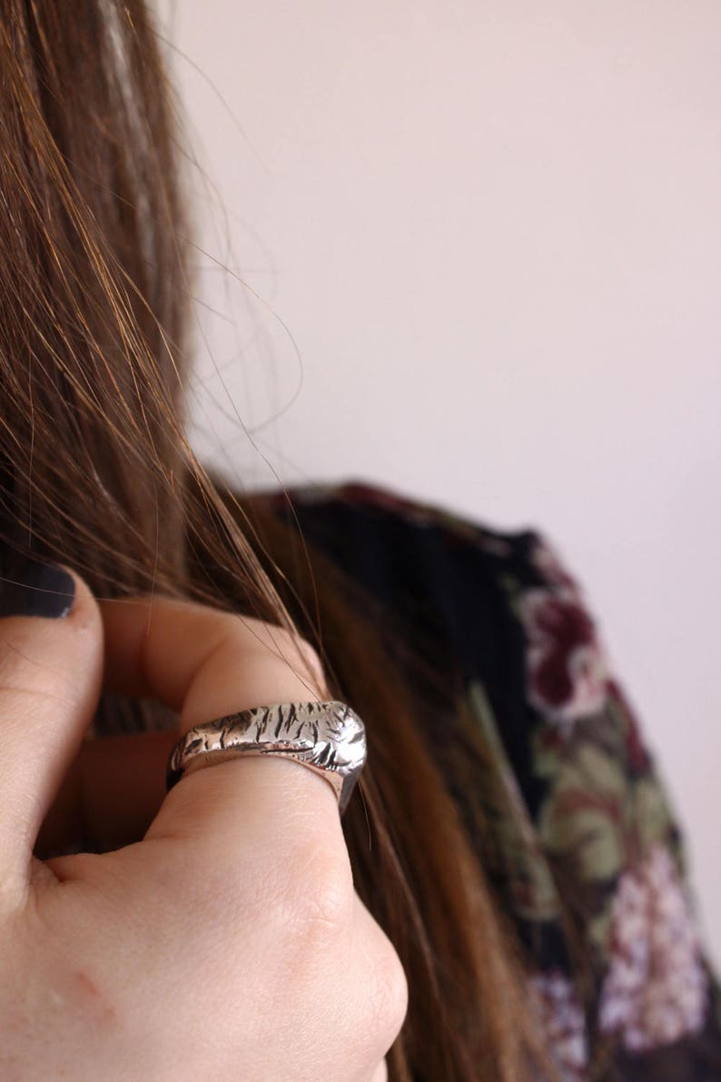 May include: A silver-toned ring featuring a textured, irregular surface with deep grooves and a polished finish. The band has a substantial, rounded profile that wraps around the finger, showcasing a rugged, organic aesthetic with visible metallic highlights.