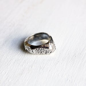 May include: A silver ring with a textured, uneven surface featuring deep, irregular grooves and a polished metallic finish. The band has a thick, organic shape with a rugged appearance, showcasing a dark, oxidized patina within the recessed areas.