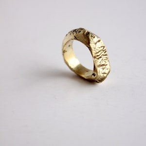 May include: A gold-colored metal ring with a textured, uneven surface. The band features a rugged, hammered appearance with visible indentations and a polished finish that reflects light across its irregular, organic-shaped outer circumference.