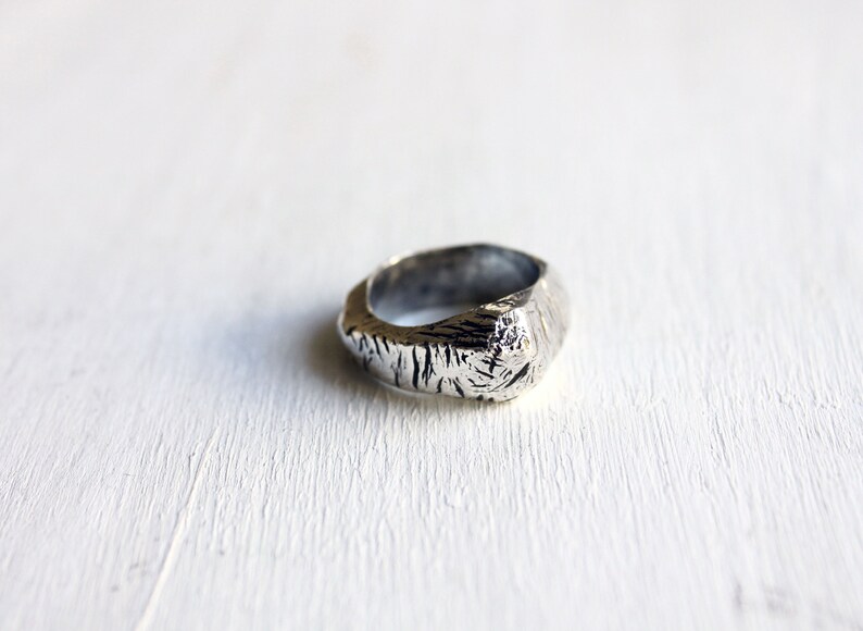 May include: A silver-toned ring with a thick, irregular band featuring a deeply textured, rugged surface. The metal has a dark, oxidized finish within the crevices, contrasting with the polished, uneven highlights across the outer circumference.