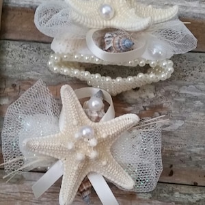 May include: Two decorative hair accessories featuring starfish, pearls, and seashells. The accessories are adorned with white tulle and ribbons. The starfish are cream-colored, and the pearls are white. The background is a weathered wooden surface.