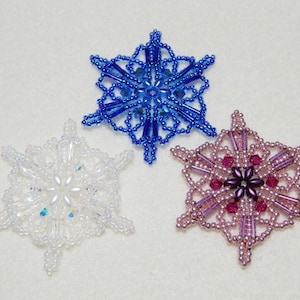 May include: Three beaded snowflake ornaments in blue, white, and pink. The blue snowflake has a crystal-like center, the white snowflake has a pearl-like center, and the pink snowflake has a purple center.