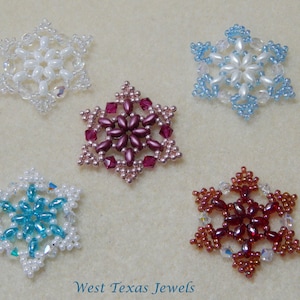 Puede incluir: Cinco adornos de copo de nieve de cuentas en varios colores, incluyendo blanco, azul, rojo y rosa. Los copos de nieve son todos del mismo tamaño y forma, y están dispuestos en un círculo sobre una superficie beige. El texto "West Texas Jewels" está impreso debajo de los copos de nieve.