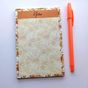 May include: A floral patterned notepad with a brown header that reads "Notes" in cursive script. The notepad is accompanied by a bright orange pen. The notepad has a floral border and a cream-colored background.