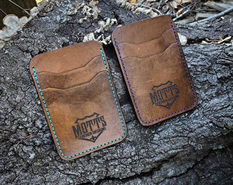 May include: Two brown leather wallets with a stitched edge. The wallets have a logo that says "Motts Leather Designs" and are on a brown wooden surface.
