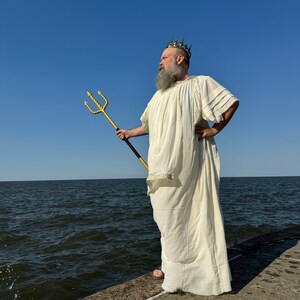 Puede incluir: Un hombre vestido con una t&uacute;nica blanca y una corona dorada sostiene un tridente dorado mientras est&aacute; de pie en un muelle de concreto con vista a una masa de agua.