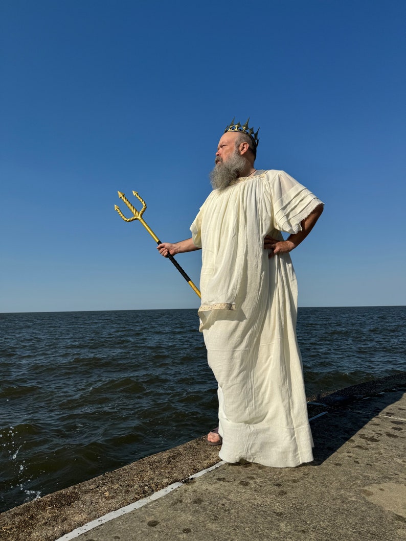 Puede incluir: Un hombre vestido con una t&uacute;nica blanca y una corona dorada sostiene un tridente dorado mientras est&aacute; de pie en un muelle de concreto con vista a una masa de agua.