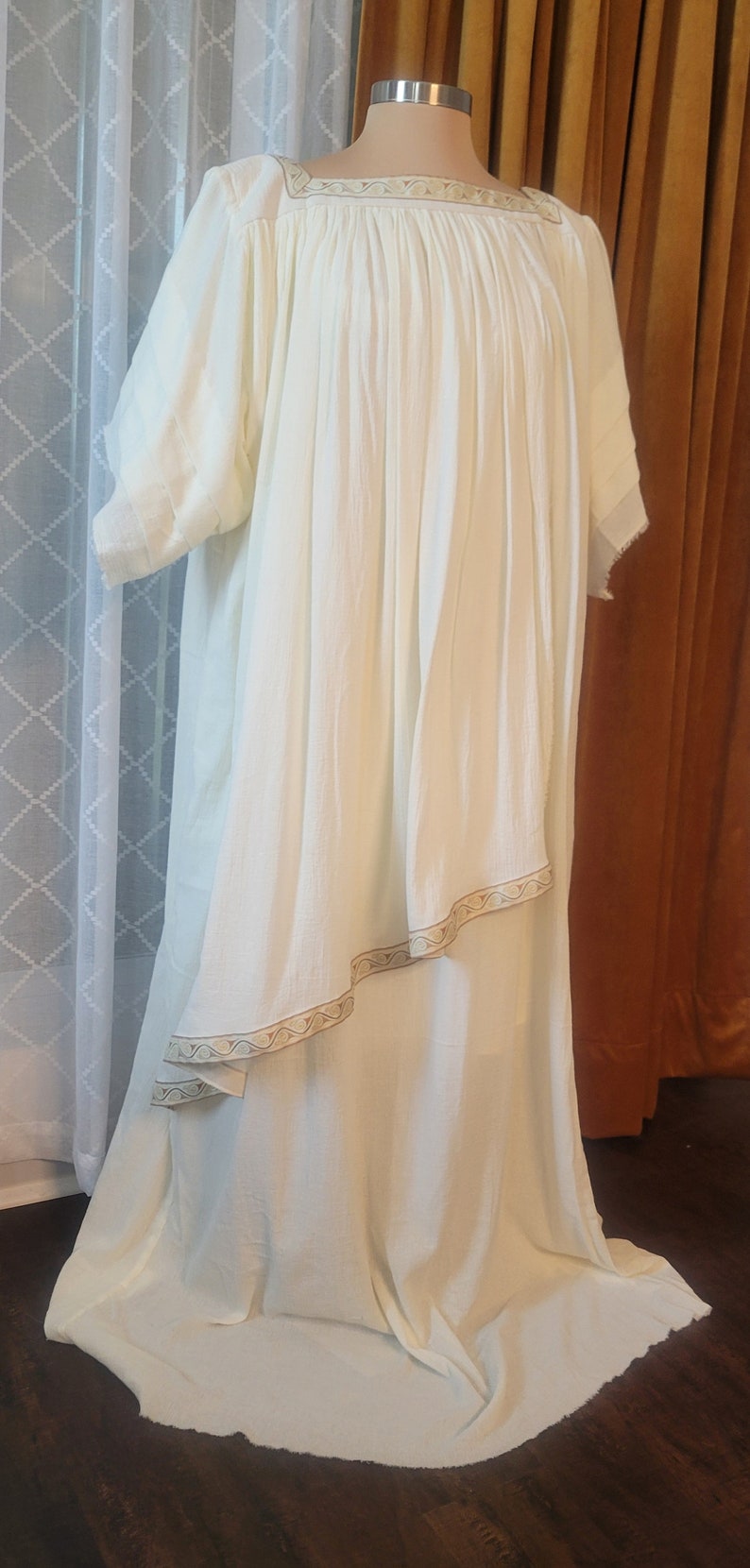 Puede incluir: Un vestido blanco fluido con un ribete decorativo alrededor del escote y el dobladillo. El vestido tiene un corte holgado y c&oacute;modo y es perfecto para una variedad de ocasiones.