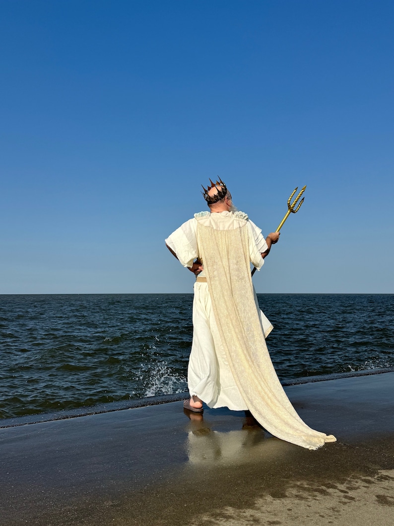 Puede incluir: Un hombre con una t&uacute;nica blanca y una corona dorada se encuentra de pie en un muelle de hormig&oacute;n con vistas a una masa de agua. Sostiene un tridente dorado en su mano derecha.
