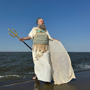 Puede incluir: Un hombre vestido como un dios del mar se encuentra de pie en un muelle de hormig&oacute;n con vistas al oc&eacute;ano. Lleva una t&uacute;nica blanca, una corona y una pieza de pecho hecha de conchas azules y blancas. Sostiene un tridente dorado en su mano derecha.