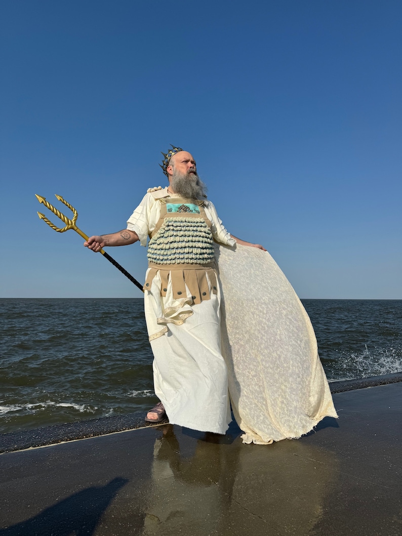 Puede incluir: Un hombre vestido como un dios del mar se encuentra de pie en un muelle de hormig&oacute;n con vistas al oc&eacute;ano. Lleva una t&uacute;nica blanca, una corona y una pieza de pecho hecha de conchas azules y blancas. Sostiene un tridente dorado en su mano derecha.
