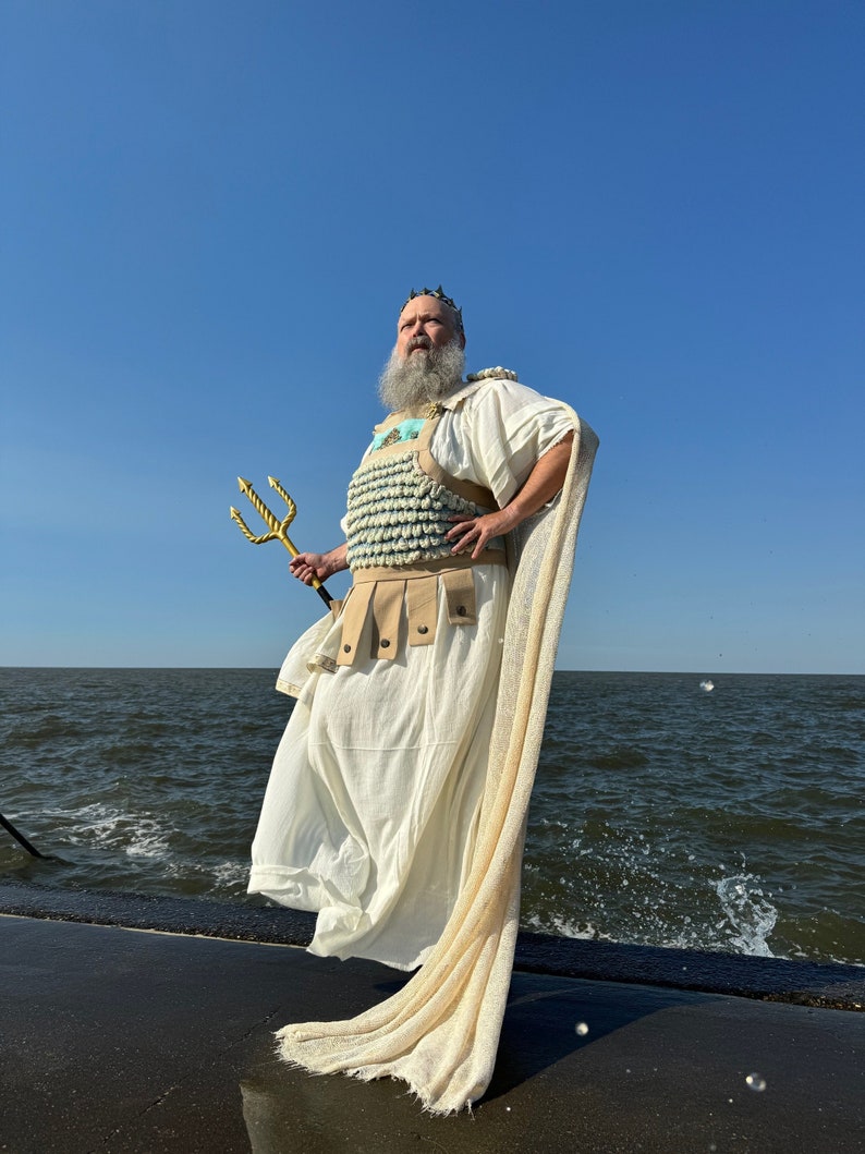 Puede incluir: Un hombre vestido como Poseid&oacute;n, el dios griego del mar, se encuentra de pie en un muelle de hormig&oacute;n. Lleva una t&uacute;nica blanca, una corona y una armadura de conchas azul y blanca. Sostiene un tridente dorado en su mano derecha. El mar est&aacute; detr&aacute;s de &eacute;l.