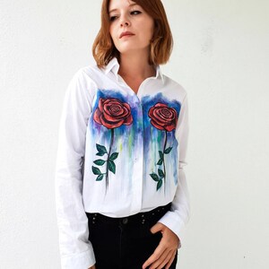 May include: White long-sleeved button-down shirt with a hand-painted design of two red roses with green stems and leaves. The roses are painted with a watercolor effect and have a blue and purple background.