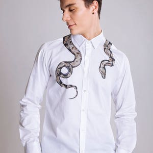 Peut inclure: Chemise blanche boutonn&eacute;e orn&eacute;e d'un motif serpent d&eacute;taill&eacute; en nuances de marron, gris et noir. Le motif serpent est imprim&eacute; sur le devant de la chemise, cr&eacute;ant un look unique. Manches longues et col classique.
