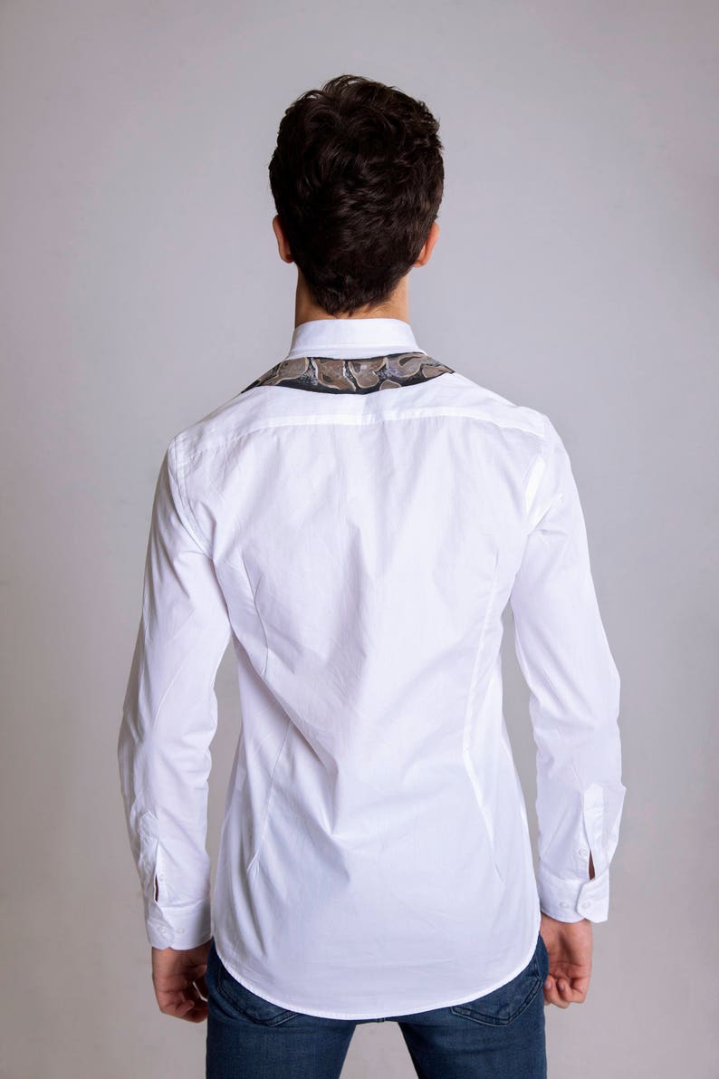 Peut inclure: Chemise blanche &agrave; manches longues avec un col &agrave; motifs. Le col pr&eacute;sente un motif dans les tons marron, gris et noir. La chemise est boutonn&eacute;e et a une coupe ajust&eacute;e, port&eacute;e avec un jean bleu.