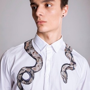 Peut inclure: Chemise blanche avec un motif de serpent peint &agrave; la main en nuances de marron, gris et noir. Le motif de serpent entoure le col et la poitrine, cr&eacute;ant un look unique et artistique. Un v&ecirc;tement de mode cr&eacute;atif et &eacute;l&eacute;gant.