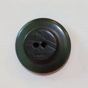 May include: A dark green button with a black, textured center and two holes. The button has a raised edge.