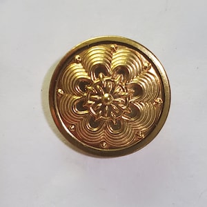 Pode incluir: Um botão redondo, dourado, com um intrincado desenho floral no centro. O botão tem um padrão texturizado em relevo com uma série de círculos concêntricos e pequenos pinos decorativos na borda.