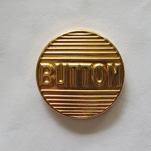 May include: A gold-colored metal button with the word "BUTTON" embossed on it. The button has a striped pattern.