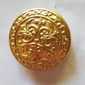May include: A gold-colored metal button with an ornate, raised design of swirling floral patterns.