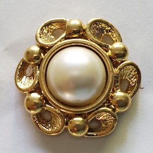 May include: A gold-toned button with a white pearl center. The button has a decorative design with intricate details.