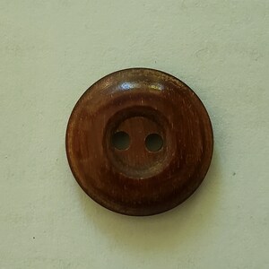 May include: A close-up of a round, wooden button with two holes. The button is a rich brown colour with a slightly glossy finish. The button's design is simple and classic, suitable for various sewing projects.