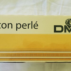 May include: A white box with the text "Coton perlé" and the DMC logo, which features a black horse head, in black text. The box has a yellow background with a thin black line border.