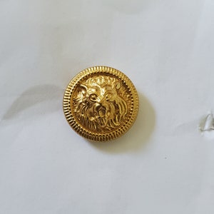 Może przedstawiać: Złoty, okrągły guzik z szczegółowym wzorem głowy lwa pośrodku. Guzik ma teksturowaną, żebrowaną obwódkę. Grzywa i rysy twarzy lwa są misternie wyrzeźbione, nadając mu królewski wygląd.