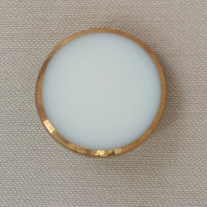May include: A round, white button with a gold rim. The button is smooth and has a glossy finish.
