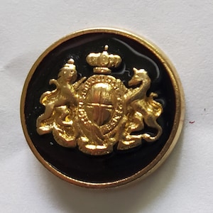 Może przedstawiać: Złoty guzik z czarnym tłem. Guzik przedstawia herb z koroną, dwoma mitycznymi stworzeniami i tarczą.