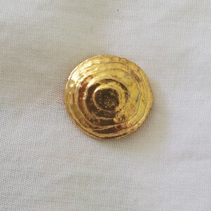 Op de afbeelding: Een gouden metalen knoop met een cirkelvormig, gelaagd ontwerp. De knoop heeft een verhoogd midden en een gladde, glanzende afwerking.