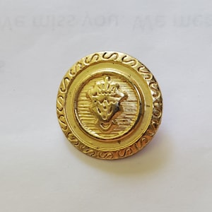 Puede incluir: Un botón de color dorado con un diseño decorativo que presenta una corona y un motivo floral estilizado.