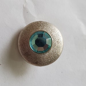Könnte beinhalten: Ein silberner Knopf mit einer strukturierten Oberfläche und einem hellblauen Strassstein in der Mitte.